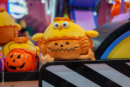 A plush toy resembling a pumpkin with a jack o' lantern face sits on a display counter, surrounded by Halloween themed items and colorful lights. photo