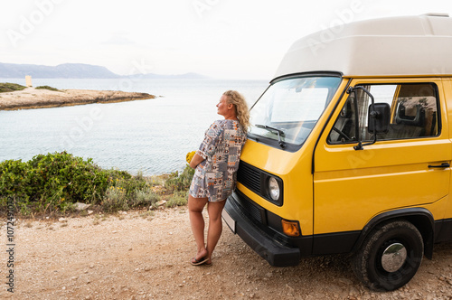Young blonde woman enjoying the sea view in her yellow retro camper van parked on a dirt road above the sea on Korcula island, Croatia