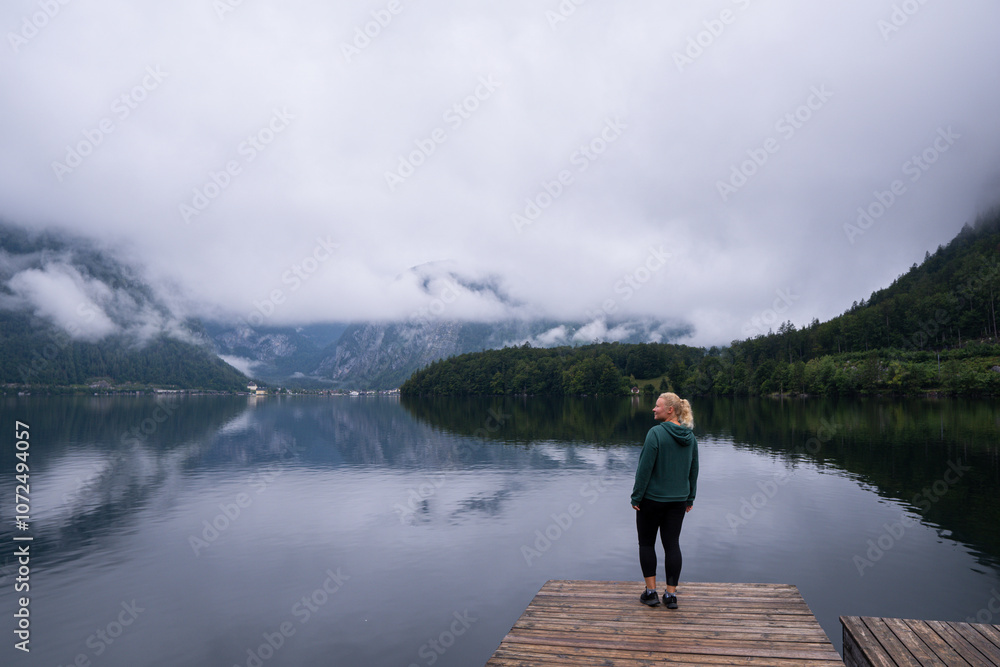 Woman standing on the Hallstatt lake shore enjoying the view of cloudy mountains, Obertraun, Austria Woman standing on the Hallstatt lake shore enjoying the view of cloudy mountains, Obertraun, Austria