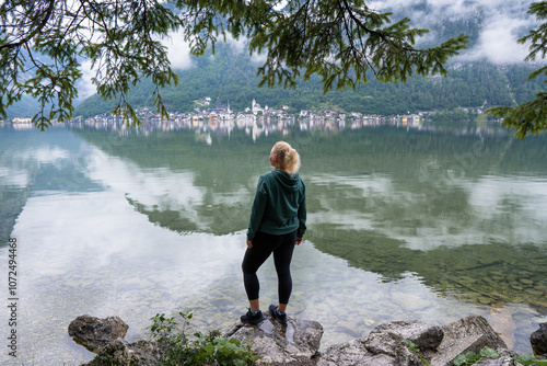 Woman standing on the lake shore enjoying the view of picturesque Hallstatt village and cloudy mountains, Obertraun, Austria