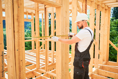 Carpenter building wooden frame house. Man measures the distance with tape measure while dressed in workwear and a helmet.