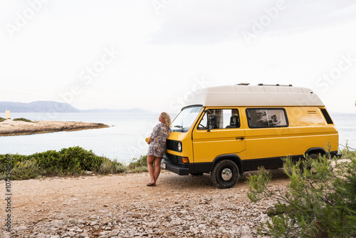 Young blonde woman enjoying the sea view in her yellow retro camper van parked on a dirt road above the sea on Korcula island, Croatia