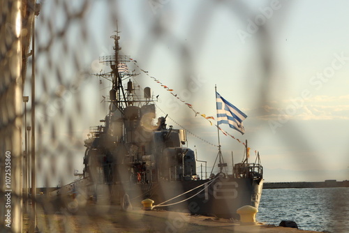 Guarded Strength: Warship and Greek Flag Through the Fence