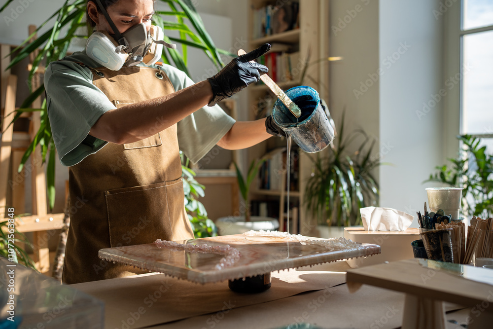Serious woman artist works with epoxy resin, creating picture in technique of liquid art. Professional female wielding mixing stick, carefully pouring fluid on to canvas. Lady fills artwork with resin Serious woman artist works with epoxy resin, creating picture in technique of liquid art. Professional female wielding mixing stick, carefully pouring fluid on to canvas. Lady fills artwork with resin