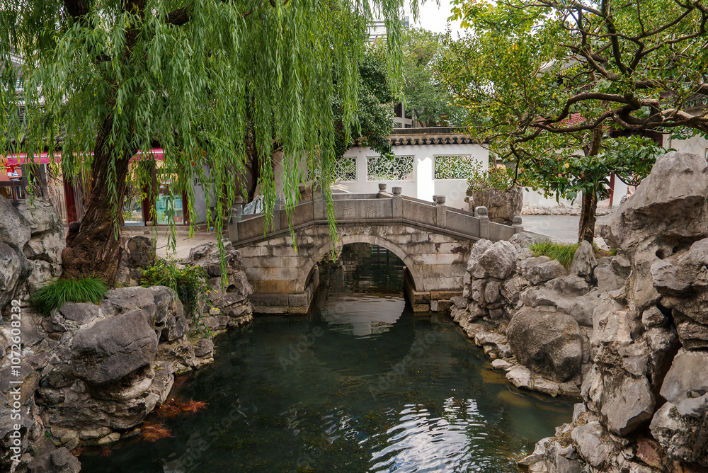 A serene garden scene in Shanghai features a stone bridge over a pond, surrounded by rock formations, lush greenery, and a willow tree. A white wall with traditional architecture is in the background. A serene garden scene in Shanghai features a stone bridge over a pond, surrounded by rock formations, lush greenery, and a willow tree. A white wall with traditional architecture is in the background.