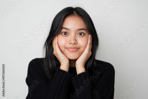 young woman with long black hair is smiling gently, resting her face on her hands against light background. Her expression conveys warmth and friendliness, making image inviting and engaging