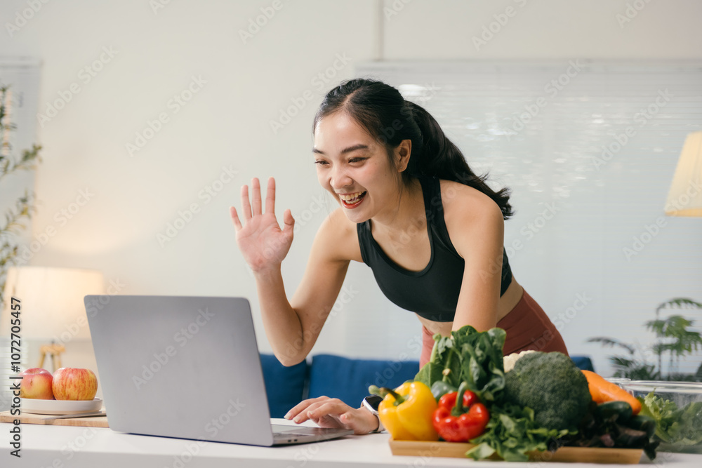 Smiling nutritionist waving at client during a video call on laptop, promoting healthy eating habits with fresh vegetables displayed on kitchen table Smiling nutritionist waving at client during a video call on laptop, promoting healthy eating habits with fresh vegetables displayed on kitchen table