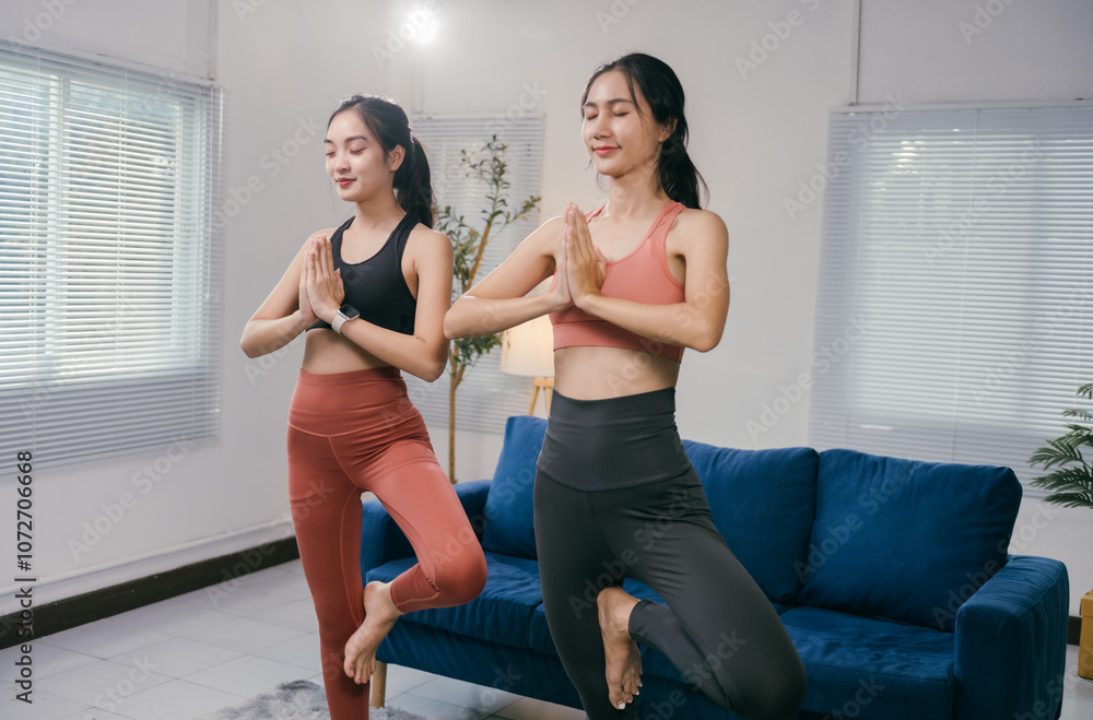 Obraz premium Two fit young women are performing the yoga tree pose in their living room, promoting balance, mindfulness, and a healthy lifestyle