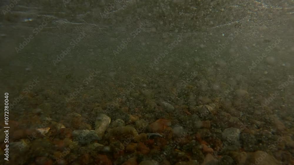 Underwater view, Powerful ocean waves rise and break on the pebble shore throwing stones at the camera lens, Split level, Slow motion Underwater view, Powerful ocean waves rise and break on the pebble shore throwing stones at the camera lens, Split level, Slow motion