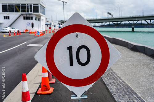 Speed Limit Sign Displaying Ten in Bold Black Numbers Surrounded by Red Circle on a Coastal Road with Cones and Bridge in Background photo