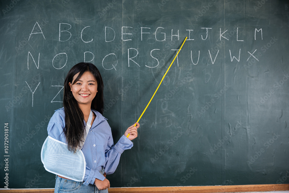 Woman is teaching class in school. She is holding yellow stick and pointing to the alphabet on the blackboard. Arm broken. Teacher confident broken arm after accident and wear arm splint for treatment Woman is teaching class in school. She is holding yellow stick and pointing to the alphabet on the blackboard. Arm broken. Teacher confident broken arm after accident and wear arm splint for treatment