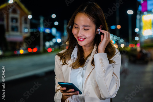 A young woman talking on her mobile phone with earphones while walking on a city street at night. Technology and wireless connection make communication easy. Handheld device in hand. photo