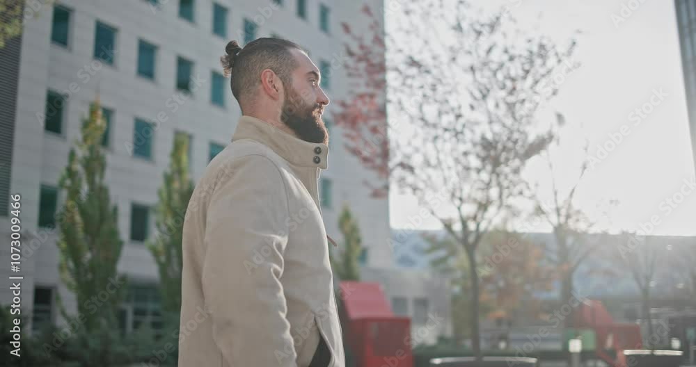 A young man with a stylish trendy hairstyle and beard in casual clothes stands on the street in the city against the background of houses and looks calmly and confidently forward A young man with a stylish trendy hairstyle and beard in casual clothes stands on the street in the city against the background of houses and looks calmly and confidently forward