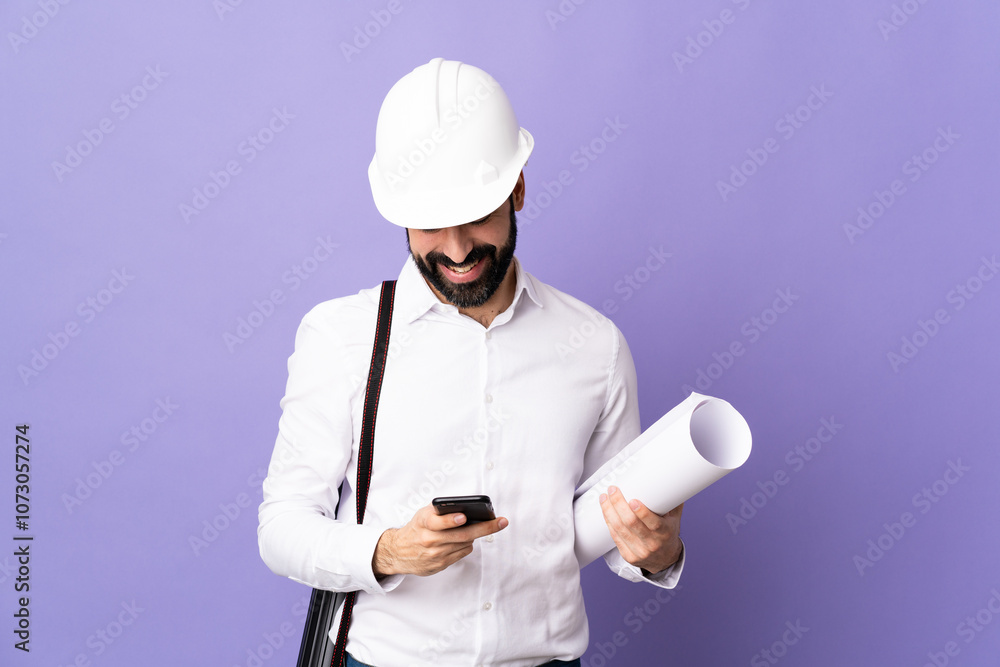 Young architect man with helmet and holding blueprints over isolated purple background sending a message with the mobile Young architect man with helmet and holding blueprints over isolated purple background sending a message with the mobile