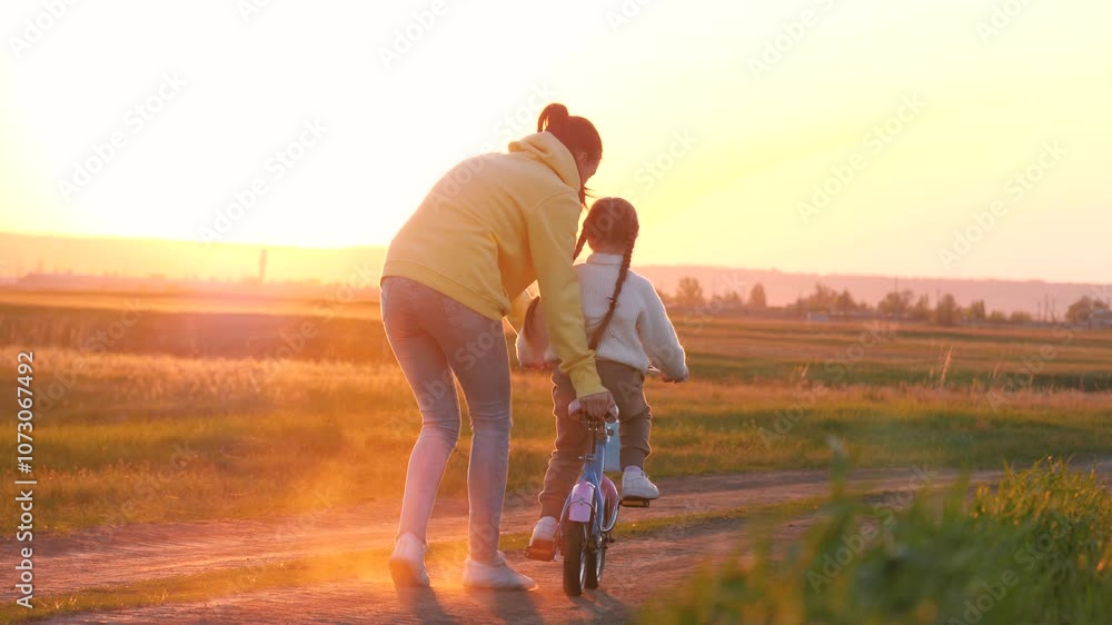Mother pulls bicycle teaching little girl to ride in sunset spring park. Little girl with mother help learns to ride bicycle in evening field. Mother and girl have fun with bicycle in twilight meadow