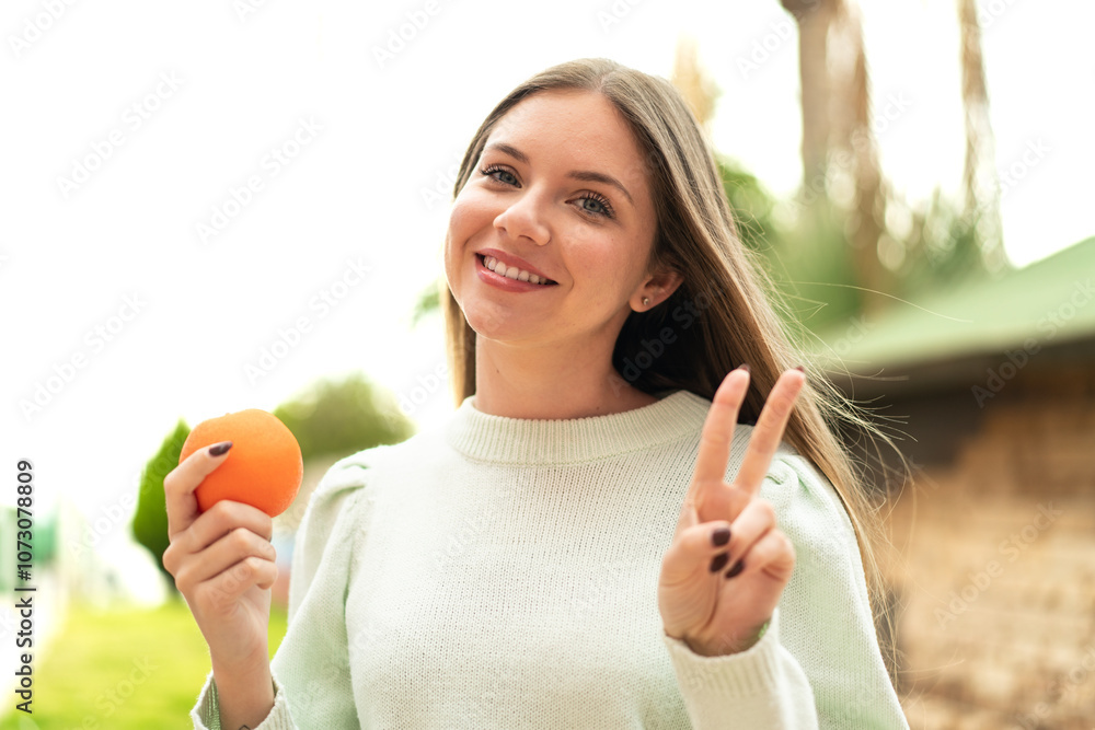 Young pretty blonde woman holding an orange at outdoors smiling and showing victory sign Young pretty blonde woman holding an orange at outdoors smiling and showing victory sign