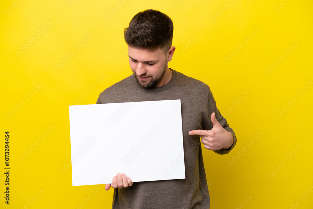Young caucasian man isolated on yellow background holding an empty placard with happy expression and pointing it Young caucasian man isolated on yellow background holding an empty placard with happy expression and pointing it