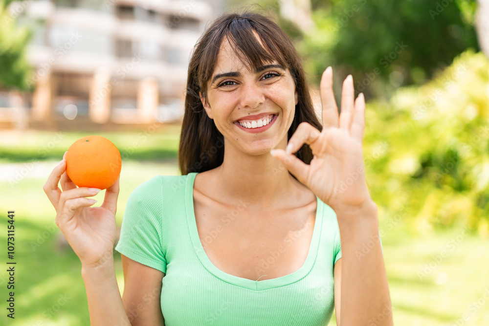 Young woman holding an orange at outdoors showing ok sign with fingers Young woman holding an orange at outdoors showing ok sign with fingers