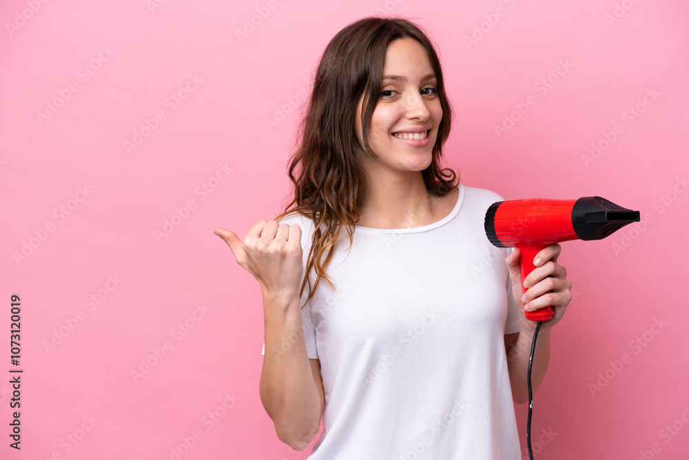 Young caucasian woman holding a hairdryer isolated on pink background pointing to the side to present a product Young caucasian woman holding a hairdryer isolated on pink background pointing to the side to present a product