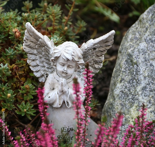 Kleiner Engel auf dem Friedhof auf einem Grab im Herbst mit Heidekraut und Grabstein