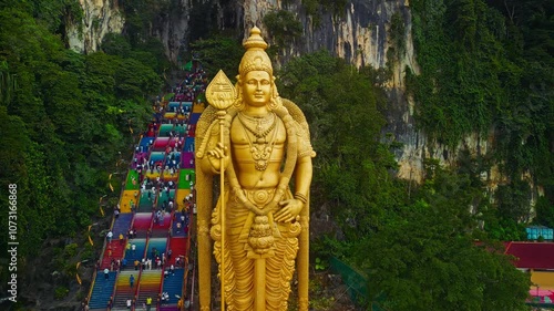 A towering golden statue of a Hindu deity stands majestically at Batu Caves in Malaysia, while numerous visitors ascend the vibrant, multicolored steps leading to the caves.