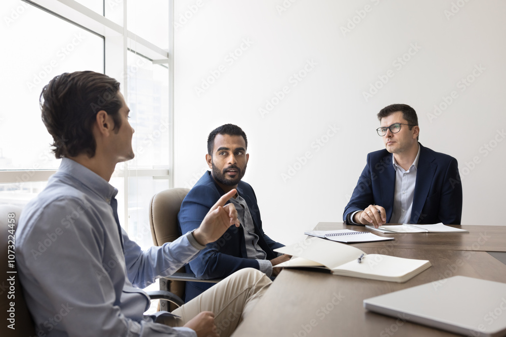 Multiethnic group of three serious young businessmen discussing work project, teamwork, management at conference table, talking in meeting room. Diverse board of directors, team of managers shot Multiethnic group of three serious young businessmen discussing work project, teamwork, management at conference table, talking in meeting room. Diverse board of directors, team of managers shot