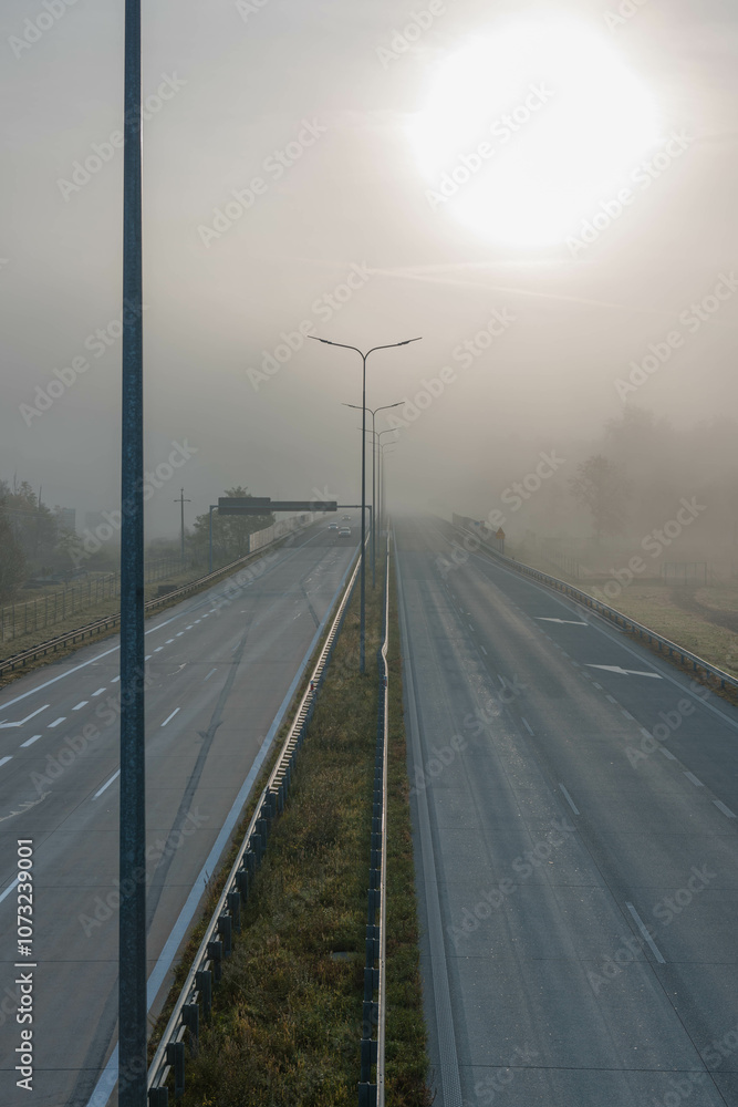 Dwujezdniowa, czteropasmowa autostrada. Nad jezdnią unosi się mgła ograniczająca widoczność. Jest ona od przodu oświetlana promieniami słońca.