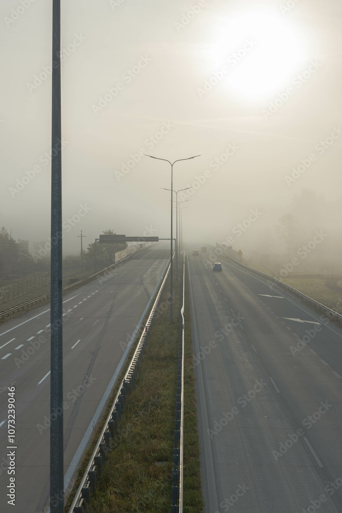 Dwujezdniowa, czteropasmowa autostrada. Nad jezdnią unosi się mgła ograniczająca widoczność. Jest ona od przodu oświetlana promieniami słońca.