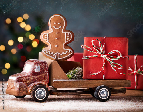 A small truck loaded with gingerbread man and  Christmas gifts on a snowy wooden table against glowing mini light bulbs creating a warm, festive background ambiance. photo