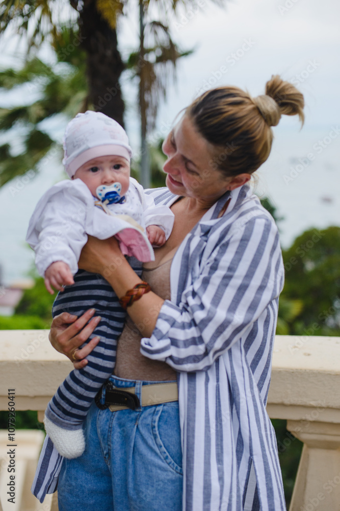 Fototapeta premium Mom and baby - little daughter in their arms in the park against the background of the sea and palm trees