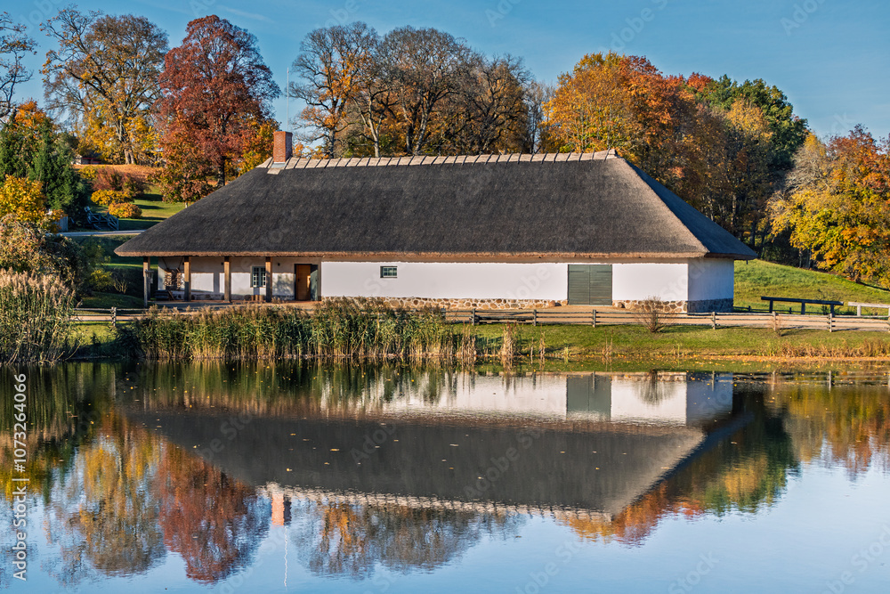 An old stone barn with a thatched roof. White building by the pond with reflection in autumn An old stone barn with a thatched roof. White building by the pond with reflection in autumn