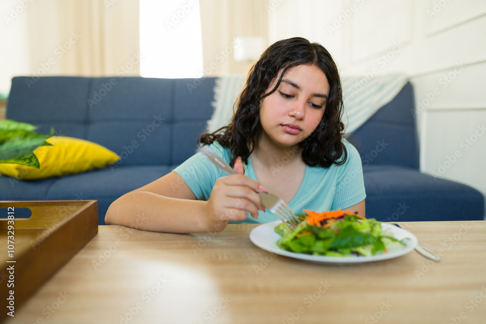Sad teenage girl reluctantly eating a healthy salad at home, struggling with eating disorder and health problems, concept of healthy diet and mental health Sad teenage girl reluctantly eating a healthy salad at home, struggling with eating disorder and health problems, concept of healthy diet and mental health