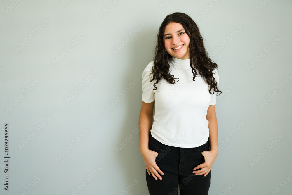 Cheerful teen girl with long curly hair in a studio portrait, wearing a white t-shirt and black pants, posing with hands in pockets against a gray background Cheerful teen girl with long curly hair in a studio portrait, wearing a white t-shirt and black pants, posing with hands in pockets against a gray background