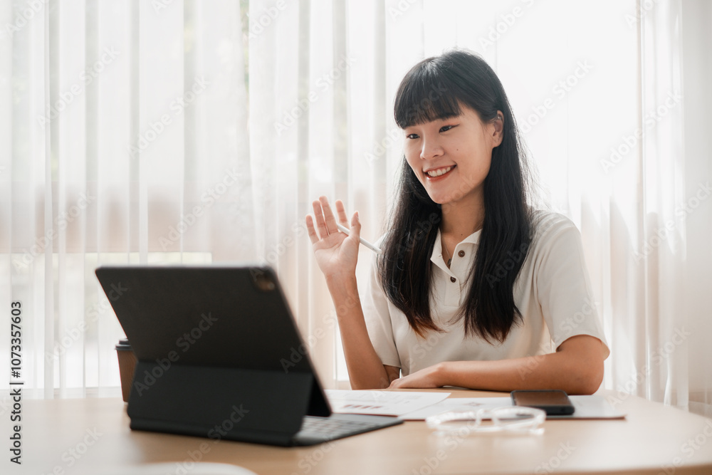 Smiling woman waving during a video call, sitting at a desk with a tablet, in a bright home office setting. Smiling woman waving during a video call, sitting at a desk with a tablet, in a bright home office setting.