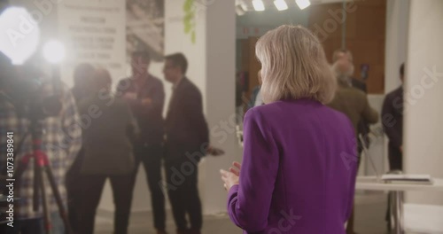 Female Reporter Speaking On-Camera. A female reporter, dressed in a purple blazer, stands in front of the camera, delivering news at a professional event.