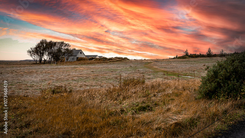 Nordic farm house with outbuildings is nestled into a small grove of trees at the far side of a grassy field showing Fall colors. It's twilight, and the clouds reflect shades of orange and yello.