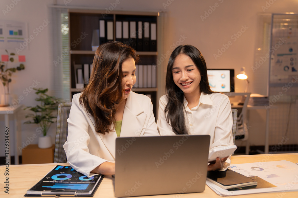 Two smiling asian businesswomen are working together using a laptop and calculating taxes in the office at night, demonstrating teamwork and dedication Two smiling asian businesswomen are working together using a laptop and calculating taxes in the office at night, demonstrating teamwork and dedication