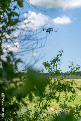 A drone flies against a bright blue sky with scattered clouds, surrounded by blurred green leaves in the foreground. aerial reconnaissance work