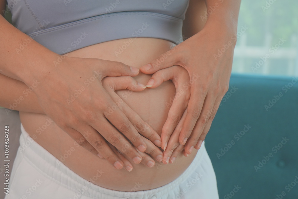 Asian husband and wife stand hugging each other, father holding hands with wife making a heart shape on the womb, wife is 7 months pregnant and happy with first child.