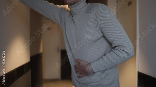 A person in a beige turtleneck clutches their stomach, appearing nauseous and distressed, in a dimly lit hallway. The subdued lighting enhances the sense of discomfort and vulnerability in the scene.