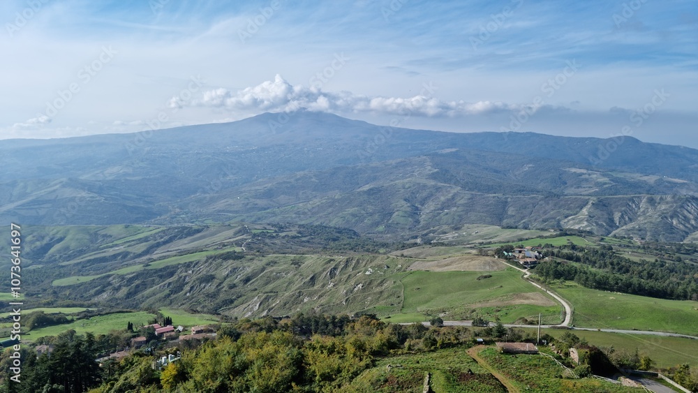 Fototapeta premium Expansive rural landscape with hills, greenery, and distant mountains under a partly cloudy sky.