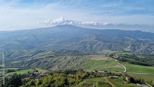 Expansive rural landscape with hills, greenery, and distant mountains under a partly cloudy sky.