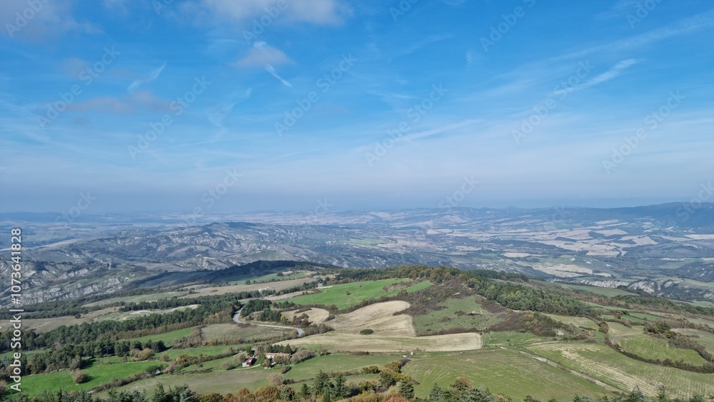 Naklejka premium Expansive rural landscape with hills, greenery, and distant mountains under a partly cloudy sky.