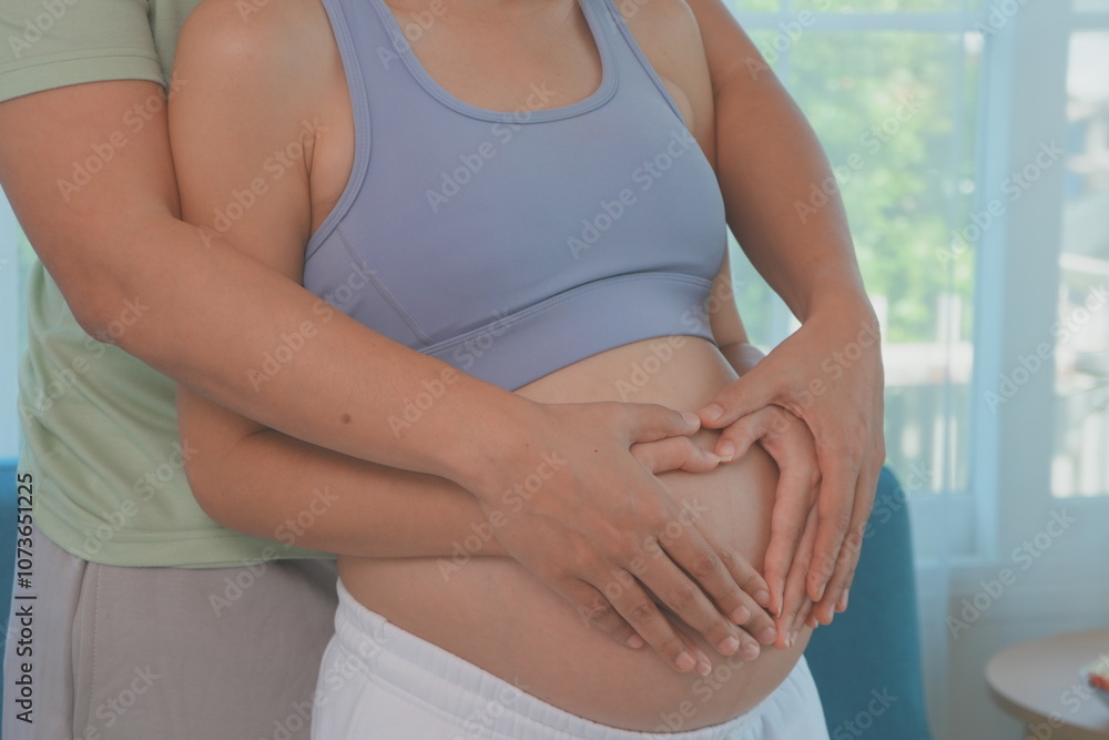 Asian husband and wife stand hugging each other, father holding hands with wife making a heart shape on the womb, wife is 7 months pregnant and happy with first child.