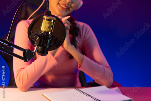 Smiling creative young beautiful Asian influencer talking in channel with positive podcast speech at night time, using laptop for presentation online at pink neon light modern studio. Stratagem. photo