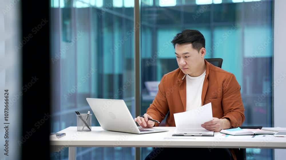 A busy asian businessman is doing paperwork using a laptop sitting in a business office. A serious male worker is working performing duties, reviewing documents, banking or making a financial report