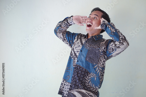 young Asian man wearing a batik shirt with a facial expression of surprise, surprise and amazement