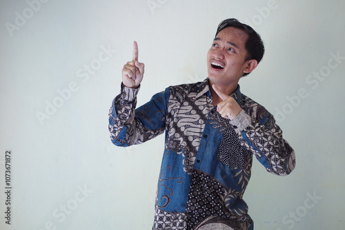 Excited young Asian man 20s wearing batik shirt pointing finger up, having great idea isolated on white background