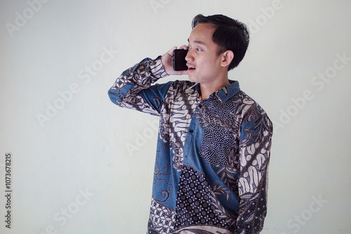 Thoughtful young asian man in a batik shirt holding a phone to his ear, looking puzzled and deep in thought against a plain background.