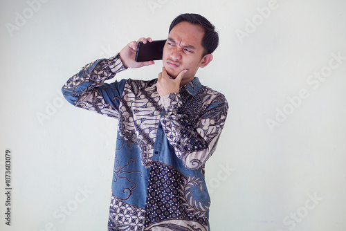 Shocked Asian man wearing batik shirt and looking at his cellphone screen, isolated on white background. expressing surprise and curiosity in a relaxed atmosphere.
